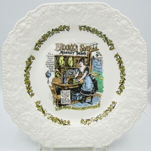 May include: A white ceramic plate with a green and gold floral border. The center of the plate features a vintage advertisement for Brooke's Soap, Monkey Brand. The ad depicts a woman washing dishes in a kitchen.