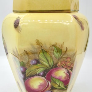 Peut inclure: Un pot à gingembre en céramique de couleur crème avec un couvercle assorti, orné d'un motif peint à la main représentant des pêches, des raisins violet foncé et des feuilles vertes. Le pot a une forme hexagonale et une finition brillante.