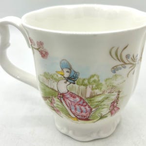 Peut inclure: Une tasse à thé en céramique blanche ornée d'une illustration décorative représentant un canard portant un bonnet bleu et un châle rose. Le décor comprend un paysage vert avec des arbres, une clôture, des fleurs roses et du feuillage vert.
