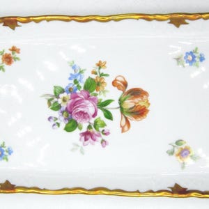 May include: An oval, white porcelain serving tray with a gold scalloped edge. The tray is decorated with colourful floral bouquets and accents. The flowers include pink, purple, orange, and blue blooms. A decorative piece for serving or display.