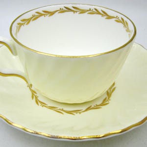 May include: A pale yellow teacup and saucer set with gold detailing and a leaf design. The cup has a curved handle and sits on a matching saucer with a scalloped edge. The gold trim highlights the rims of the cup and saucer.