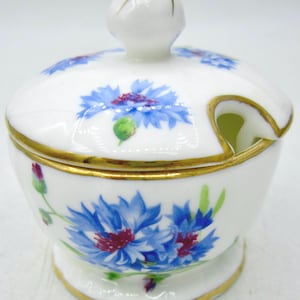 May include: White ceramic sugar bowl with a lid, decorated with hand-painted blue cornflower floral designs and gold trim. The bowl has a small footed base and a spoon rest. The lid has a small knob for easy handling.