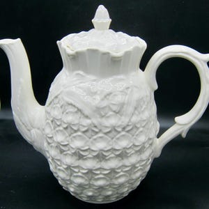 May include: White ceramic teapot with a floral design. The teapot has a lid with a knob on top and a handle on the side.