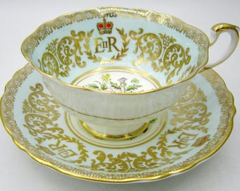 Könnte beinhalten: Ein Vintage-Tee-Set aus feinem Porzellan. Die Tasse und Untertasse sind hellblau mit goldenen Verzierungen und Verzierungen. Die Tasse zeigt eine Krone und die Buchstaben "ER". Die Untertasse hat einen gewellten Rand.