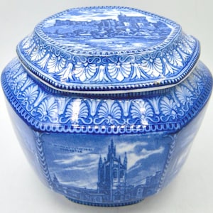 May include: A blue and white ceramic jar with a matching lid, featuring detailed transferware illustrations of architectural landmarks. The body displays the text St. Nicholas Cathedral Newcastle, while the lid shows a castle scene with decorative borders.