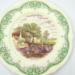 May include: A decorative plate with a pastoral scene featuring a river, trees, and a horse-drawn cart. The plate has a cream-colored background with a green floral border and the text "THE HAY-WAIN After Constable".
