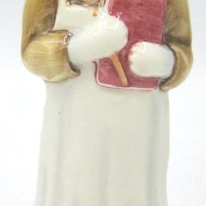 Peut inclure: Une figurine en céramique représentant un chien anthropomorphe à tête noire, vêtu d'une veste beige et d'un tablier blanc. La figurine tient un livre rouge et un petit crayon beige dans ses pattes, sur une base circulaire à motif de pierre.