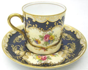 May include: A vintage teacup and saucer set. The cup and saucer feature a navy blue and cream color scheme with gold accents. Floral designs and ornate patterns decorate the set. The cup has a gold handle and a white interior.