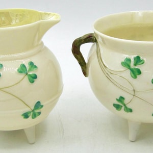 May include: Cream ceramic creamer and sugar bowl set. The creamer has a spout and handle, the sugar bowl has two handles. Both feature green shamrock designs and stand on three small legs. A charming kitchenware set.