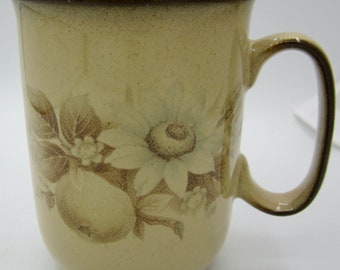May include: A beige ceramic mug with a brown rim and handle. The mug features a floral design with brown and white sunflowers, leaves, and fruit.