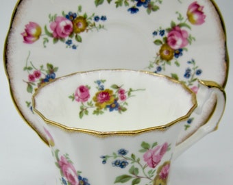 May include: A white teacup and saucer set with a floral pattern featuring pink roses, blue accents, and gold trim. The teacup has a scalloped edge and a handle. The saucer is round with a matching floral design.