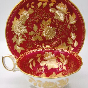 May include: A red and gold teacup and saucer set. The cup and saucer feature a floral design with gold accents. The rim of the saucer and the cup are trimmed with a gold geometric pattern. The cup has a white base with gold floral details.