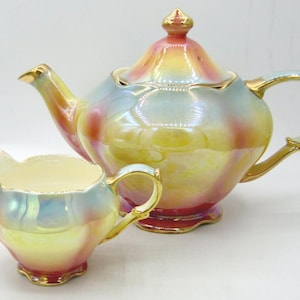 Peut inclure: Un service à thé vintage comprenant une théière et un pot à crème. L'ensemble présente une finition nacrée avec un mélange de couleurs, dont le jaune, le rose et le bleu. Des bordures dorées ornent les bords et les poignées.