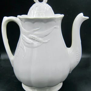 May include: A white ceramic teapot with a lid, handle, and spout. The teapot features embossed wheat stalks and a decorative handle. The lid has a small loop for easy removal. The teapot is designed for serving tea or coffee.