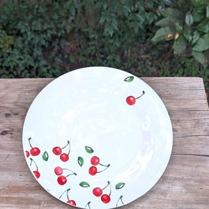 Może przedstawiać: Biały ceramiczny talerz z ręcznie malowanym wzorem wiśni. Talerz ma czerwone wiśnie z zielonymi liśćmi rozrzuconymi wokół krawędzi. Talerz jest ustawiony na drewnianej powierzchni.