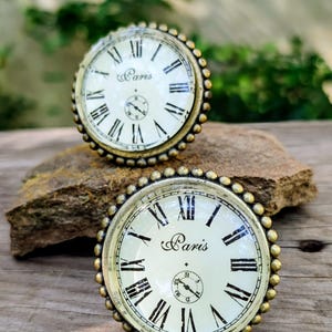 May include: Two antique-style clock faces with Roman numerals and the word "Paris" printed on the cream-colored face. The clock faces have a gold-colored beaded border and are set on a stone.