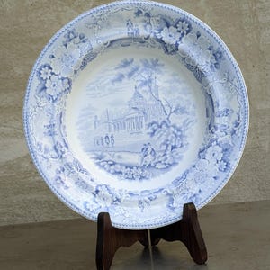 May include: A blue and white antique plate with a scenic design. The plate features a central landscape with a building and figures, surrounded by floral patterns. The plate is displayed on a wooden stand.