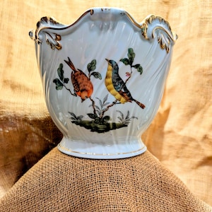 May include: A white ceramic planter with a scalloped rim and gold accents. The planter features a hand-painted design of two birds perched on a branch with green leaves.