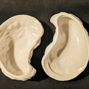 Puede incluir: Dos platos de cerámica blanca con forma de concha de ostra. Los platos están vacíos y tienen un acabado liso y brillante.