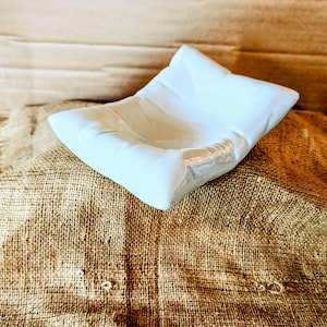 May include: White ceramic dish with a unique, folded design. The dish is sitting on a burlap surface.