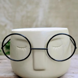 Puede incluir: Una maceta de cerámica color crema con forma de rostro con ojos cerrados y una nariz pequeña. Gafas de montura metálica negra están adheridas a la maceta. La maceta está sobre una superficie de madera.
