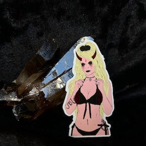 May include: A sticker featuring a cartoon illustration of a person with blonde hair, red horns, and dark makeup, wearing a black bikini. A crystal is in the background.