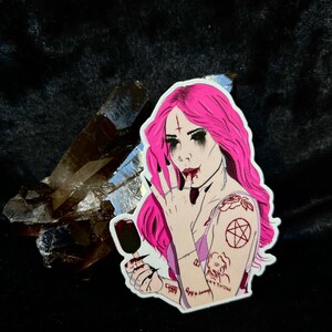 Può includere: Un adesivo con un'illustrazione gotica di una donna con capelli rosa, trucco scuro e tatuaggi, che tiene in mano un ghiacciolo. L'adesivo ha un bordo bianco ed è posto su uno sfondo scuro con un cristallo.