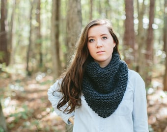 May include: A woman with long brown hair wearing a light blue button-down shirt and dark blue jeans. She is wearing a dark gray knitted scarf around her neck. The woman is standing in a forest with trees in the background.