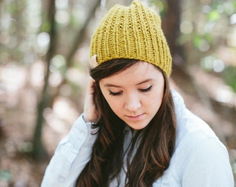 May include: A woman with long brown hair is wearing a light blue button-down shirt and a yellow knitted beanie.