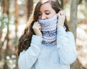 May include: A woman wearing a light blue denim shirt and a gray and white knit cowl scarf. The scarf is pulled up over the lower face. The background is a blurred forest scene with sunlight filtering through the trees.