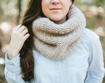 May include: A woman wearing a light blue shirt with a beige knitted cowl neck scarf. The scarf has a herringbone pattern.