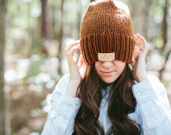 May include: A brown knit beanie with a small rectangular leather patch featuring a mountain design. The beanie is worn low, partially obscuring the face. The background is a blurred forest scene.