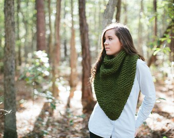 May include: A woman wearing a white shirt and a green knitted scarf stands in a forest. The scarf is triangular in shape and has a textured knit.