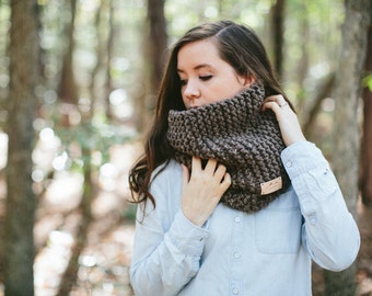May include: A woman wearing a brown knitted infinity scarf with a leather tag. The scarf is thick and cozy, perfect for cold weather.