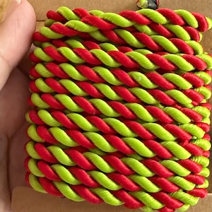 May include: A spool of twisted cord featuring a vibrant two-tone design with alternating strands of bright lime green and saturated cherry red. The cord is neatly wrapped around a rectangular brown cardboard backing for organized storage and display.