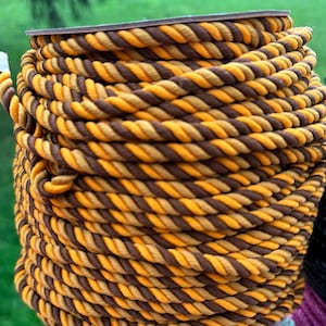 May include: A tightly wound coil of rope featuring alternating strands of brown and golden yellow. The rope is thick and appears to be made of a durable material, suitable for various crafting or decorative purposes.