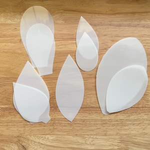 May include: Six white paper templates for crafting, shaped like leaves and teardrops, on a wooden surface.