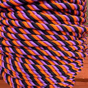 May include: A coiled rope featuring alternating strands of black, orange, and purple. The rope is tightly twisted, creating a textured surface. The color scheme suggests a Halloween or festive theme.