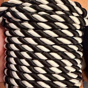 May include: A spool of thick, twisted cord featuring a repeating pattern of alternating black and white strands. The cord is wound tightly in horizontal rows around a flat, light brown cardboard backing, creating a uniform, textured appearance.