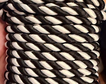 May include: A spool of thick, twisted cord featuring a repeating pattern of alternating black and white strands. The cord is wound tightly in horizontal rows around a flat, light brown cardboard backing, creating a uniform, textured appearance.