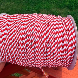 May include: A large spool of twisted cord featuring a repeating diagonal pattern of bright red and white stripes. The cord is wound tightly around a cylindrical core, creating a continuous texture of alternating colors across the entire surface.