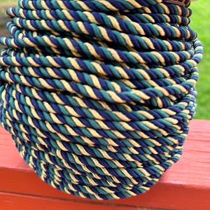 May include: A spool of twisted cord featuring a repeating pattern of dark blue, teal, and metallic gold threads. The cord is wound tightly into a cylindrical shape, showing a consistent braided texture throughout the entire length of the spool.