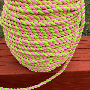 May include: A large spool of twisted rope in vibrant neon pink and green. The rope is tightly wound, creating a textured surface. The spool sits on a wooden surface, with the rope extending outwards.