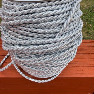 May include: A spool of silver twisted rope. The rope is tightly wound and has a shiny, metallic appearance. The spool is made of cardboard and is sitting on a wooden surface.