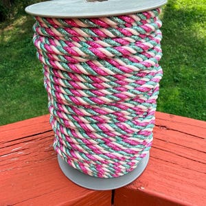 May include: A spool of thick, twisted rope featuring a repeating pattern of bright pink, light pink, and shimmering teal green strands. The rope is wound tightly around a gray plastic cylindrical spool with flat circular ends at the top and bottom.