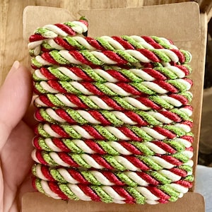 May include: A spool of decorative twisted cord featuring a repeating pattern of metallic red, metallic green, and white strands. The cord is wound tightly around a rectangular cardboard bobbin, showcasing a multi-colored, textured rope design.