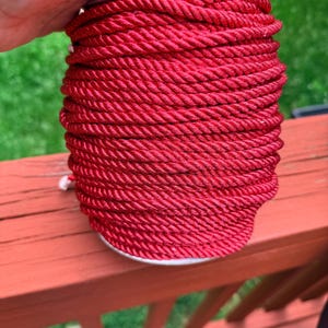 May include: A spool of bright red rope. The rope is tightly wound around a cardboard core. The texture of the rope is visible, with each strand clearly defined. The spool is resting on a red wooden surface.