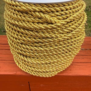 May include: A spool of gold-colored rope. The rope is tightly wound and has a twisted texture. The spool has a white top and is sitting on a red-orange surface. The rope appears to be made of a shiny, metallic material.