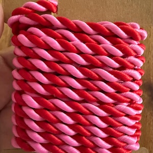 May include: A length of twisted cord featuring alternating strands of bright red and light pink satin-like material. The cord is neatly wound around a rectangular brown cardboard spool, displaying a consistent braided texture throughout the length.