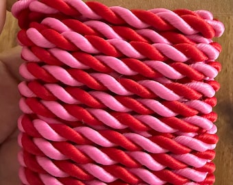 May include: A length of twisted cord featuring alternating strands of bright red and light pink satin-like material. The cord is neatly wound around a rectangular brown cardboard spool, displaying a consistent braided texture throughout the length.